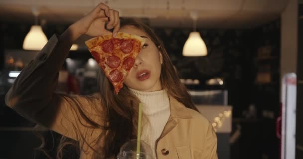 감각적 인 젊은 금발 소녀가 카페에서 칵테일을 들고 서서 얼굴 일부를 덮고 있는 페퍼로니 피자 한 조각을 들고 있습니다 — 비디오