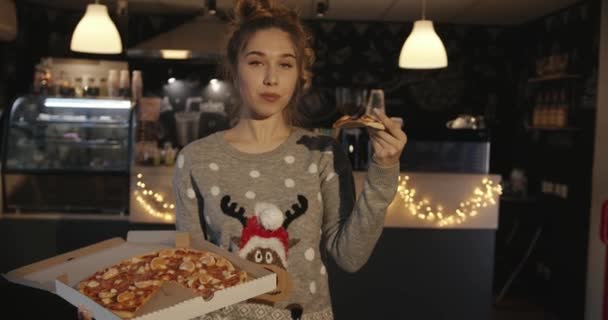 Een charmant meisje in een kersttrui staat in een café en eet pizza met mandarijnse plakjes uit een doos. Langzame beweging — Stockvideo