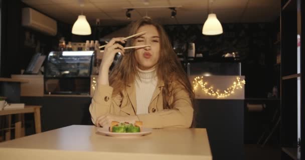 En glad ung flicka med långt blont hår sitter på ett café vid ett bord med en tallrik sushi och håller ätpinnar nära ansiktet — Stockvideo