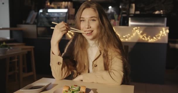 Atrakcyjna młoda dziewczyna z luźnymi długimi włosami siedzi w kawiarni przy stole z talerzem sushi z sosem i trzyma pałeczki w zębach — Wideo stockowe