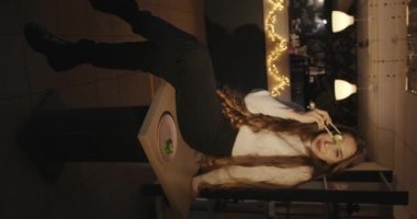 Uzun sarı saçlı şehvetli genç kız bir kafede bir masada oturuyor ve göz hizasında yeşil havyarda yemek çubukları suşisi tutuyor. Dikey video