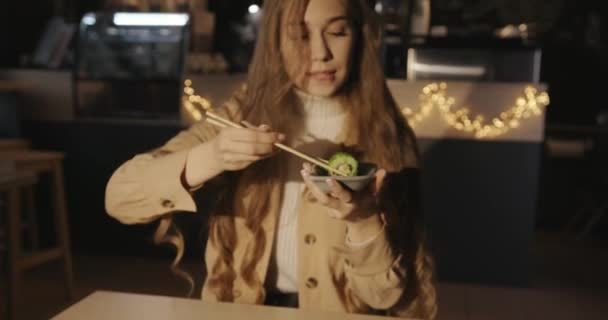 Seorang gadis muda yang cantik duduk di meja di sebuah kafe dan makan sushi dengan kaviar ikan terbang hijau, mencelupkannya ke dalam saus — Stok Video