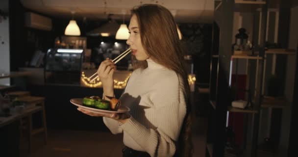 Delikatna młoda dziewczyna w białym swetrze trzyma talerz sushi w dłoniach i smakuje bułeczkami z czerwoną rybką siedząc na stole w kawiarni — Wideo stockowe