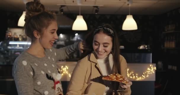 Deux jeunes filles aux cheveux longs se tiennent dans un café et mangent la pizza d'une boîte en riant. Mouvement lent — Video