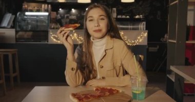 Uzun sarı saçlı güzel bir kız bir kafede oturur ve lezzetli pepperonili pizza ve ferahlatıcı bir içecek yemekten hoşlanır.