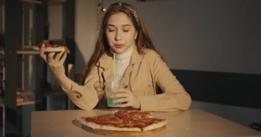 Beyaz kazaklı ve bej ceketli tatlı genç kız bir kafede pizza yiyor ve pipetle limonlu mavi bir kokteyl içiyor.