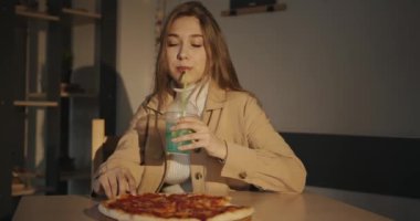 Kafede sarı saçlı narin bir genç kız pipetle kokteyl içer ve elinde bir dilim pizza tutar.