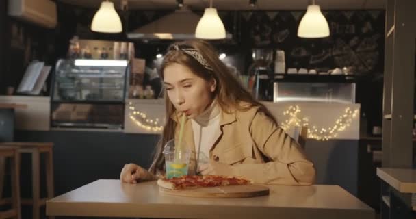 Una hermosa rubia joven se sienta en un café en una mesa con rebanadas de pizza en una tabla de madera y bebe un cóctel azul con limón a través de una paja — Vídeos de Stock