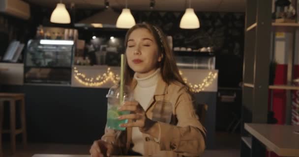 Piękna młoda dziewczyna w beżowej kurtce jedząca pizzę w kawiarni pijąca koktajl przez słomkę i mrugająca uśmiechnięta — Wideo stockowe