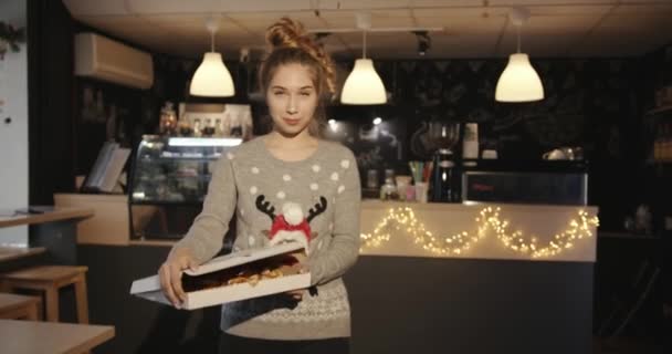 Una linda jovencita en un suéter con un ciervo navideño camina por el café y abre una caja de pizza. Movimiento lento — Vídeo de stock