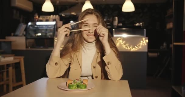 Menina bonita em uma jaqueta bege senta-se em um café em uma mesa com um prato de sushi e grimaces clicando pauzinhos — Vídeo de Stock