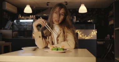 Uzun sarı saçlı şehvetli genç kız bir kafede bir masaya oturur ve yemek çubuklarıyla geleneksel Japon suşisi yer.