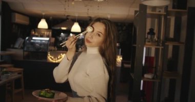 Güzel, genç, uzun saçlı bir kız elinde bir tabak suşiyle bir kafede bir masada oturuyor ve elinde yemek çubuklarıyla göz hizasında bir rulo tutuyor.