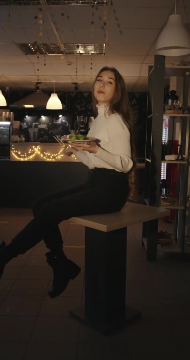 En charmig ung flicka med långt blont hår sitter på ett bord i ett café med en tallrik sushi i händerna och dinglar med benen. Vertikal video — Stockvideo