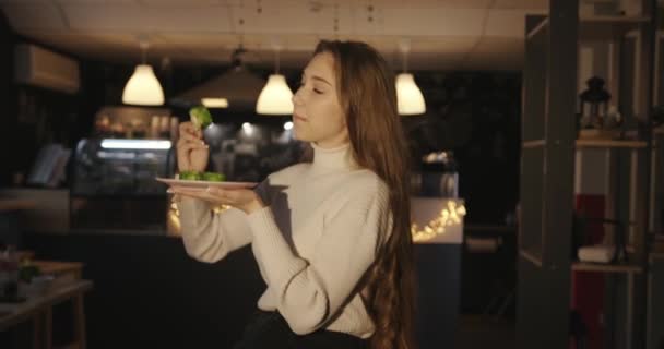 Een leuk jong meisje in een witte trui zit in een café en eet met eetstokjes sushi in groene vliegende viskaviaar. Zijaanzicht — Stockvideo