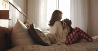 Ev kıyafetleri ve terlikleriyle sevimli genç bir kız güneş ışığında bir yatakta oturuyor ve kollarında komik bir köpekle ona sarılıyor.