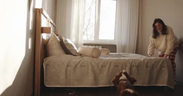 Une jeune fille joyeuse joue avec son chien assis sur le lit sur le fond de la fenêtre et souffle dans son museau — Video