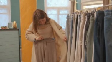 Kotlu ve kazaklı genç bir kız elbise mağazasından alışveriş yapıyor ve pantolonlarını ona iliştirerek pantolon deniyor.