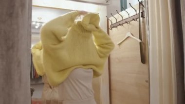 Sevimli genç kız bir giyim mağazasının soyunma odasında seçtiği kazağını giyer ve saçını düzeltirken güler.