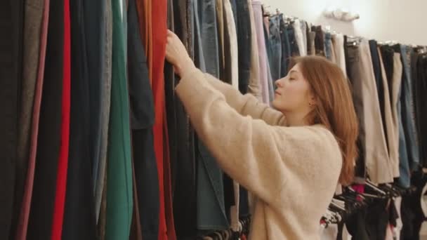 Hermosa chica joven de compras y clasificación a través de la ropa en el estante en una gran tienda — Vídeos de Stock