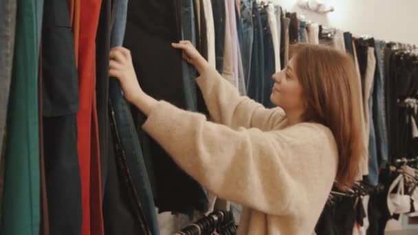 Smiling young girl chooses clothes in a fashion store and goes through models on the rack. Slow motion — Stock Video