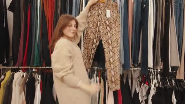 Ładna młoda kobieta robi zakupy w sklepie odzieżowym i zdejmuje spodnie z półki bada je — Wideo stockowe