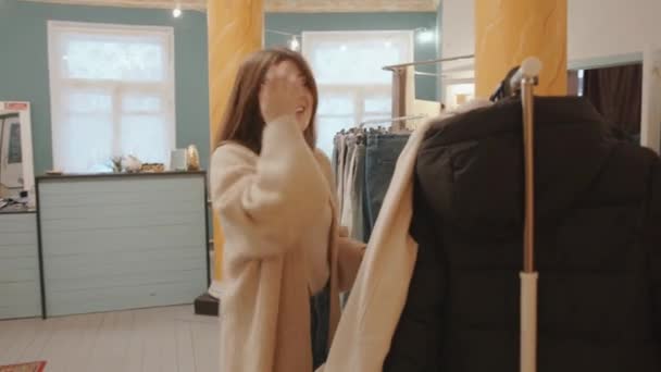 Aantrekkelijk jong meisje kiest kleding in een stijlvolle boetiek en onderzoekt een jas op een hanger en glimlacht — Stockvideo