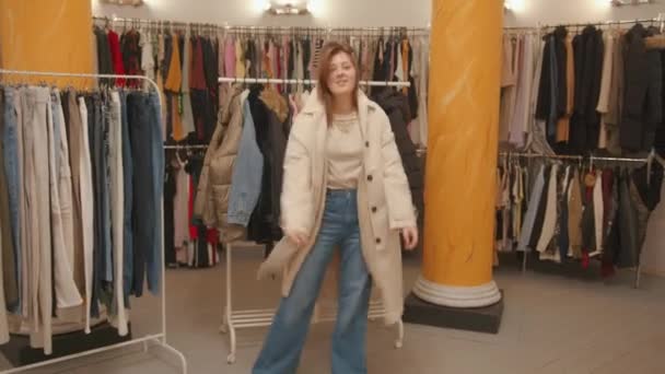 Elegante chica joven en pantalones vaqueros hipster está de compras en una tienda de ropa y está girando alrededor de probar un nuevo abrigo — Vídeos de Stock