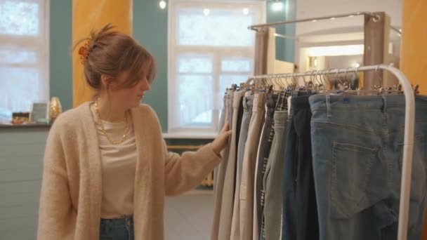 Egy csinos fiatal lány elsétál a ruha sorok mellett a boltban, és megvizsgálja a kabátot egy vállfán. Lassú mozgás. — Stock videók