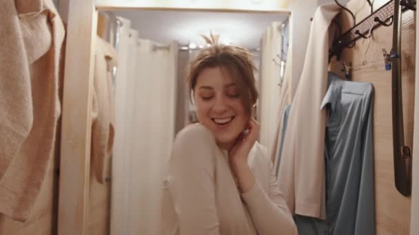 En öm ung flicka står i ett provrum i en klädaffär mot bakgrund av en spegel och poserar leende mot kameran — Stockvideo
