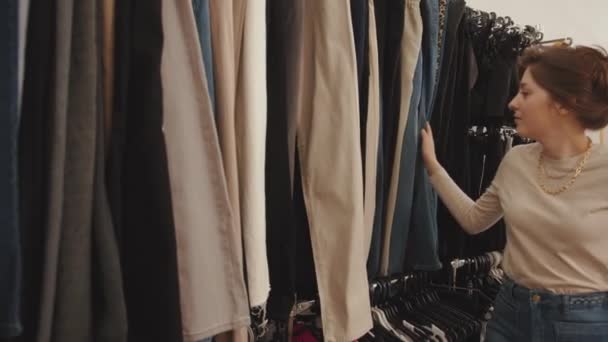 Милая молодая девушка во время шопинга в бутике проходит мимо стойки с одеждой и выбирает модный наряд для себя — стоковое видео