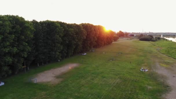 Blick von oben auf ein grünes Naherholungsgebiet in der Nähe der Bäume mit ruhenden Menschen am Flussufer vor dem Hintergrund des Sonnenuntergangs — Stockvideo