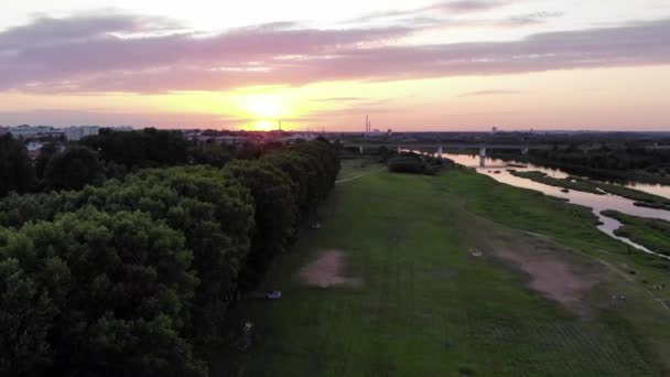 Дронна зйомка зеленої зони відпочинку на березі річки та міських будинків на тлі заходу сонця — стокове відео