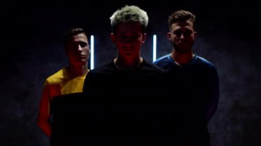 Genç futbolcular, karanlık bir stüdyoda duvarın arkasında, üzerinde neon lambaların çizgileri olan bir duvarın arkasında duruyor ve içlerinden biri elinde bir futbol topu tutuyor. 4K