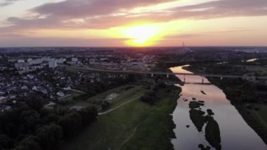 Nehir kıyısında, ortasında yeşil adalar ve gün batımının arka planında bir yol köprüsü olan bir şehrin çarpıcı insansız hava aracı görüntüleri.