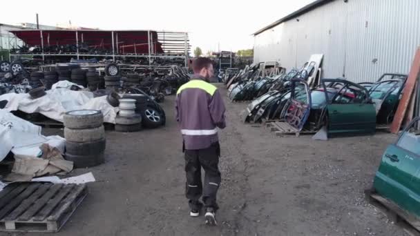 Een jonge bebaarde automonteur met bril loopt tussen de rijen van reserveonderdelen in de openlucht en maakt aantekeningen in een notitieboekje. Schieten vanuit een helikopter — Stockvideo