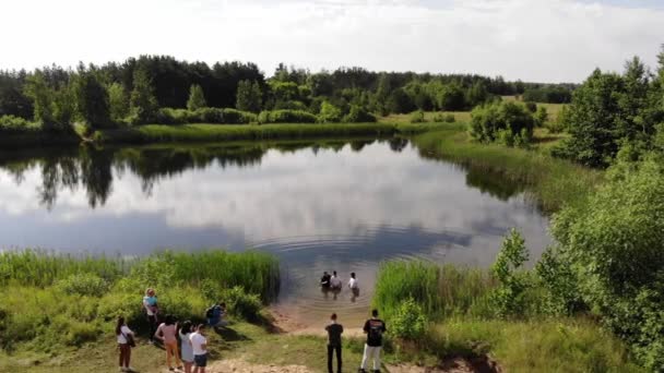 Misk, Bielorrusia - 15 de agosto de 2020: Dos pastores bautizan a un joven en nombre de Cristo en un lago en un día de verano frente a un grupo de amigos en la orilla. Disparos con drones — Vídeo de stock