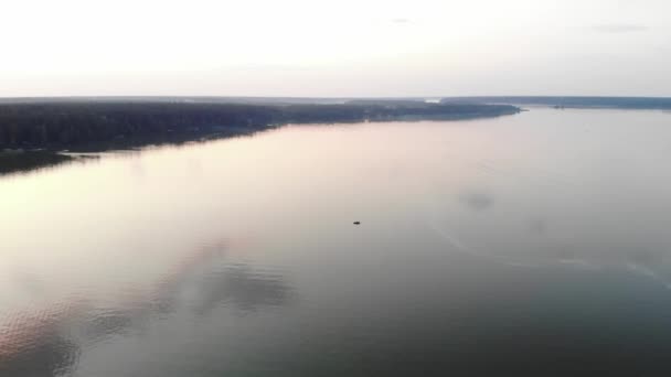 Veduta aerea di un pescatore in barca nel mezzo di un grande lago tra foreste e nuvole rosa riflesse nell'acqua al tramonto — Video Stock