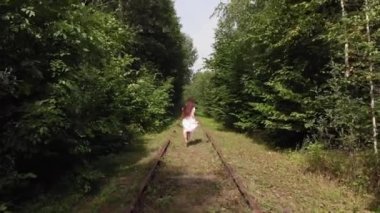 Ormanda terk edilmiş bir demiryolunda çıplak ayakla koşan beyaz elbiseli, uzun saçlı genç bir kızın hava manzarası.