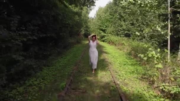 白いドレスを着た美しい少女は、森の中の古い放棄された鉄道に沿って裸足で走っています。空中風景 — ストック動画