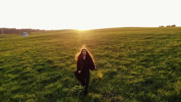 Luchtfoto van een jong meisje in een zwarte trench jas loopt op een groene weide met uitwaaierend haar tegen een zonsondergang achtergrond — Stockvideo