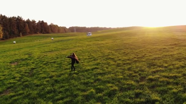 Aerial view of a young girl spinning with arms outstretched in the sunset on a large green field. Happiness concept — Stock Video