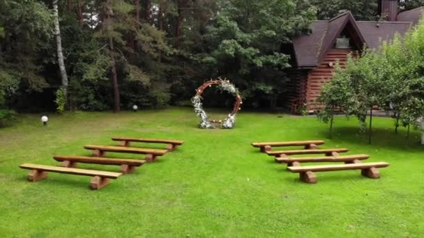 Drohnenaufnahmen eines Blumenbogens und Sitzreihen in der Nähe eines Holzhauses für eine Trauung im Freien — Stockvideo