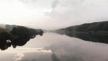 Büyük bir nehrin üzerindeki sisli hava manzarası kıyılarda ağaçlar ve su yüzeyinde bulutların yansıması.