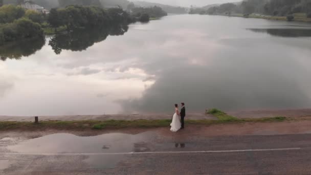 Panna młoda i pan młody stoją twarzą w twarz na tamie na tle rzeki i mgły nad lasem. Widok z lotu ptaka — Wideo stockowe