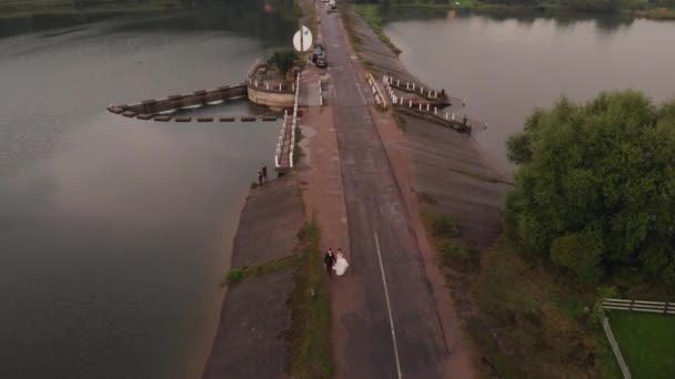 Drohnenaufnahmen des Brautpaares, das auf dem Damm über den Fluss entlang der Straße läuft, bei einem Fotoshooting — Stockvideo