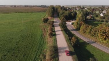 Yeşil ağaçlar ve tarlalarla çevrili bir köyün yakınındaki toprak yolda bir arabanın hava görüntüsü.