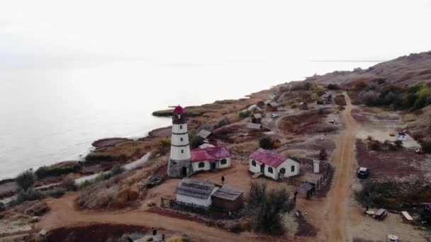 Luchtfoto van een vuurtoren op een heuvel met gebouwen op de binnenplaats en de kronkelende kust van de zee — Stockvideo