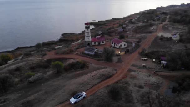 Vista aérea de un coche que conduce a lo largo de la carretera cerca del faro en una colina junto al mar — Vídeos de Stock