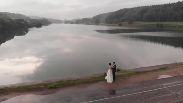 Nevěsta a ženich stojí tváří v tvář na přehradě na pozadí řeky a mlhy nad lesem. Letecký pohled — Stock video